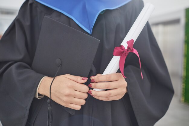 Ritratto ritagliato di giovane donna irriconoscibile in possesso di diploma e berretto di laurea mentre si trova all'aperto durante la cerimonia, copia spazio