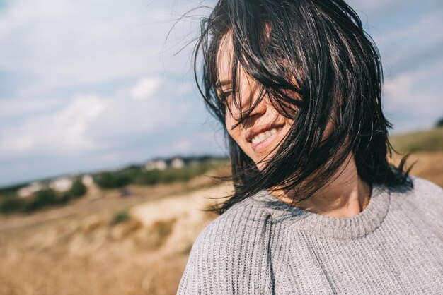 Ritratto ritagliato orizzontale di bella donna sorridente che indossa un maglione che è giocoso con i capelli ventosi e spensierato in posa sul cielo del sole e sullo sfondo della natura Persone di viaggio e concetto di stile di vita