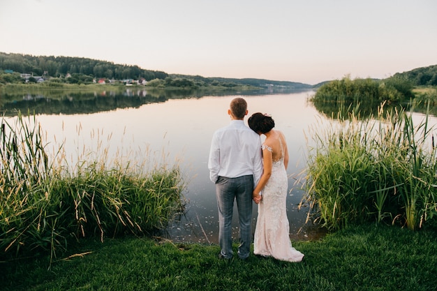 Ritratto romantico da dietro delle coppie di nozze che stanno insieme sulla riva al tramonto e che godono della vista del lago.