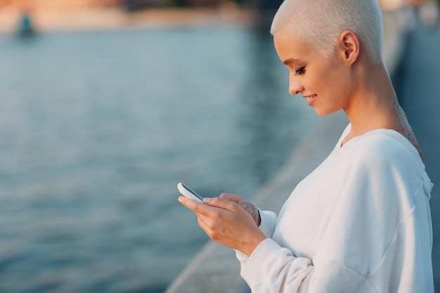 Ritratto sorridente all'aperto dei capelli corti biondi della giovane donna millenario con il cellulare