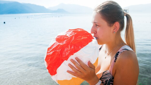 Ritratto tonificato di giovane madre sorridente che soffia aria in pallone da spiaggia gonfiabile sul mare.