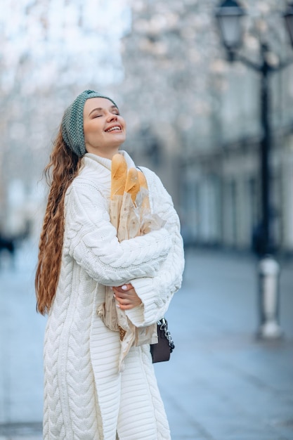 Ritratto urbano alla moda di sfondo sfocato con una giovane donna attraente in abito bianco che stringe 2 mani attorno a un pacchetto di pane. Occhi chiusi e un sorriso raggiante. Pubblicità di prodotti da forno.