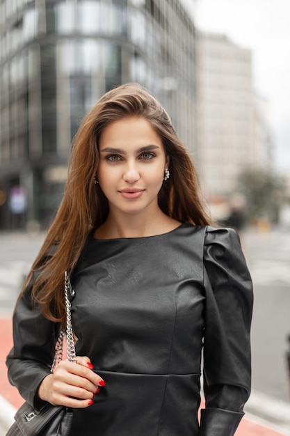 Ritratto urbano femminile di una bella giovane donna d'affari di successo in abiti neri di moda con un vestito e una borsa di pelle che cammina nella città moderna