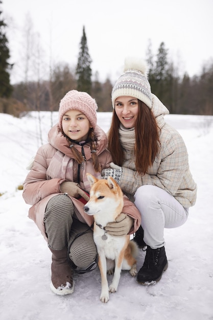 Ritratto verticale a figura intera della moderna madre e figlia felici in posa con un cane e guardando la telecamera mentre si gode una passeggiata all'aperto nella foresta invernale