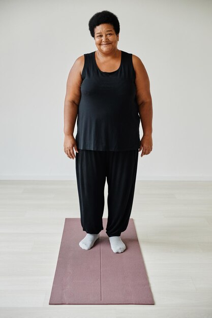 Ritratto verticale a figura intera di donna anziana sorridente in piedi sul tappetino da yoga mentre si lavora al chiuso