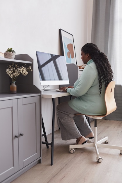 Ritratto verticale a figura intera di una donna formosa afroamericana che utilizza il PC alla scrivania e mentre si gode il lavoro da casa in interni minimi