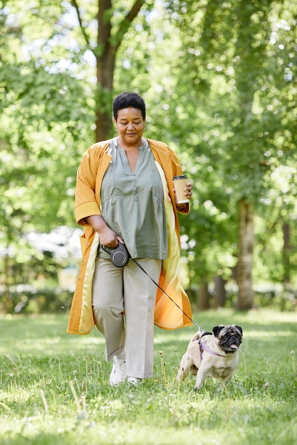 Ritratto verticale a figura intera di una donna nera sorridente che cammina con un cane carino nel parco e si gode il caffè