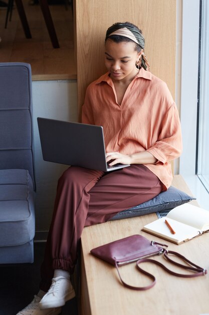 Ritratto verticale a figura intera di una giovane donna d'affari afroamericana che utilizza il computer portatile mentre è seduta alla finestra in un bar o in ufficio e si gode il lavoro