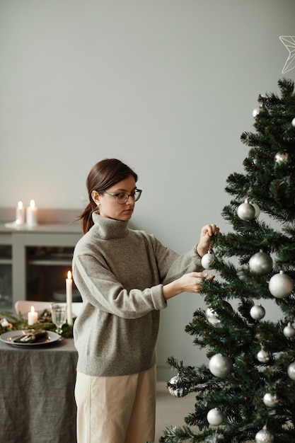 Ritratto verticale a vita alta di giovane donna che decora l'albero di Natale a casa in elegante argento e gr...