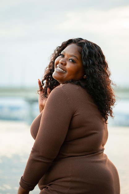 Ritratto verticale del primo piano della donna della pelle scura dell'afroamericano grassoccio sorridente riccio che osserva la posa della macchina fotografica contro il cielo blu