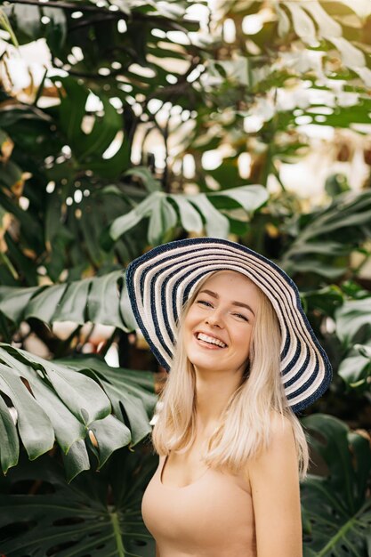 Ritratto verticale del primo piano di giovane donna caucasica attraente felice con capelli biondi, cappello d'uso, costume da bagno, sorridente con il sole che gode, rilassandosi alla località di soggiorno tropicale. concetto di estate.