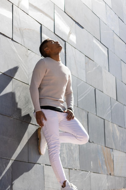 Ritratto verticale dell'uomo latino vestito di bianco appoggiato a un muro grigio che guarda in alto con gli occhi chiusi nel pensiero.