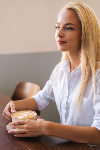 Ritratto verticale della giovane donna bionda attraente che tiene tazza di caffè caldo del latte con il bello modello che si siede al tavolo di legno