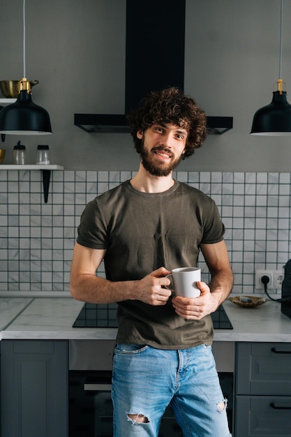 Ritratto verticale di allegro giovane barbuto che tiene in mano una tazza con il caffè del mattino guardando la fotocamera in piedi in cucina