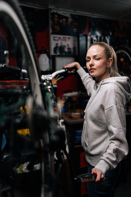 Ritratto verticale di attraente meccanico di bici femminile che ripara e fissa la bicicletta di montagna in piedi sul portabiciclette in officina di riparazione bici con interni scuri