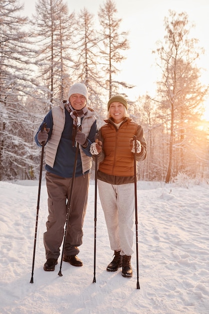 Ritratto verticale di coppia senior attiva che si gode la camminata nordica con i pali nella foresta invernale e looki