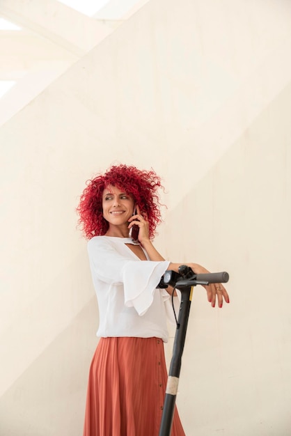 Ritratto verticale di donna d'affari latina sorridente con capelli afro rossi che parla sullo smartphone mentre si appoggia la mano su uno scooter elettrico