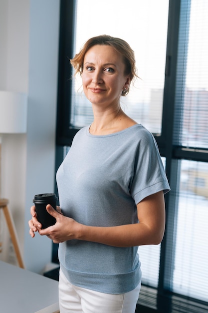 Ritratto verticale di donna positiva che tiene una tazza di caffè e guarda la fotocamera in piedi sullo sfondo di una grande finestra in ufficio