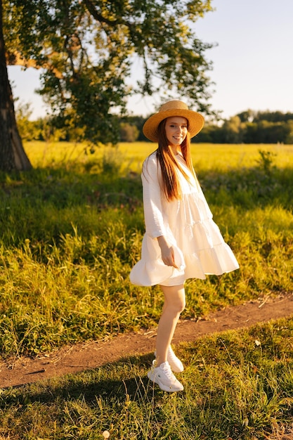 Ritratto verticale di giovane donna dai capelli rossi sorridente in cappello di paglia e abito bianco in piedi in posa guardando la fotocamera sul bellissimo campo di erba verde