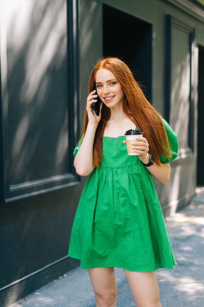 Ritratto verticale di giovane donna sorridente in abito verde che tiene una tazza di caffè da asporto, parlando al telefono cellulare camminando per la strada della città nella soleggiata giornata estiva, guardando la fotocamera.