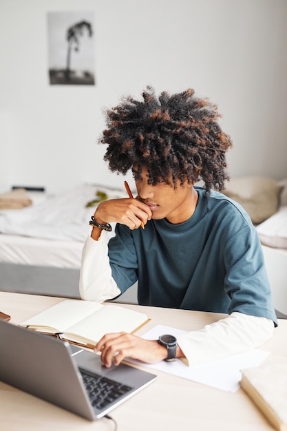 Ritratto verticale di un adolescente afroamericano che usa il portatile mentre studia a casa o nel dormitorio del college
