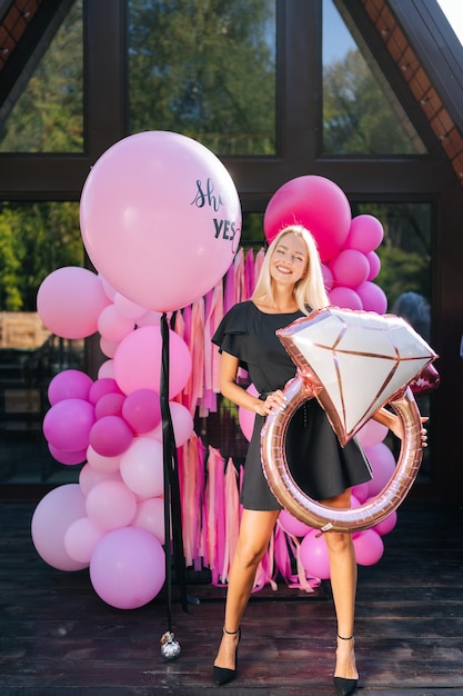 Ritratto verticale di un'elegante donna bionda in abito estivo che posa con vari palloncini di elio colorati e festivi in piedi sullo sfondo di una casa di campagna sorridendo guardando la telecamera