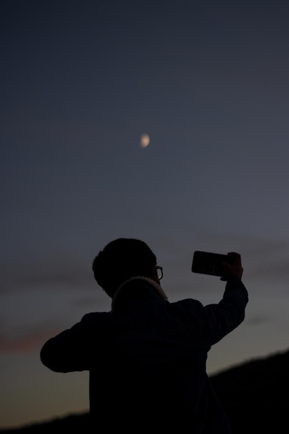 Ritratto verticale di un giovane che scatta una foto della Luna con il suo cellulare al tramonto al tramonto