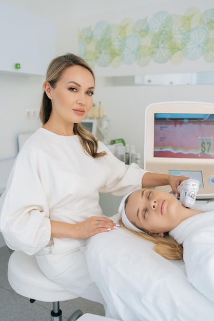 Ritratto verticale di un medico estetista che utilizza macchine laser e ultrasuoni SMAS per ringiovanire la pelle di una donna sdraiata su un divano medico in una clinica di medicina estetica che guarda la telecamera