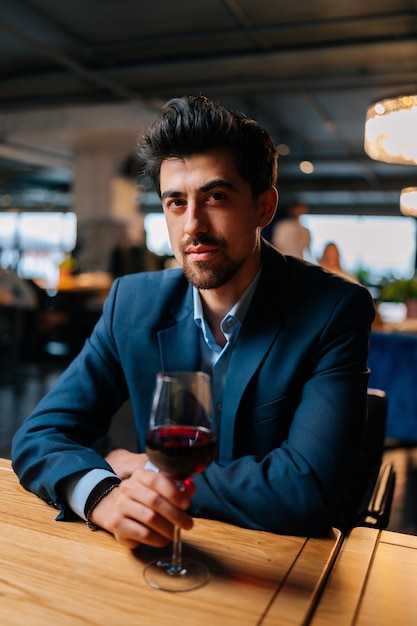 Ritratto verticale di un uomo elegante e sicuro in abito di moda che tiene bicchieri di vino rosso seduto al tavolo in un ristorante con interni scuri che guarda l'obbiettivo