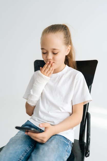 Ritratto verticale di una bambina bionda carina con il braccio rotto avvolto in un bendaggio di gesso bianco che guarda lo schermo del telefono cellulare seduto alla sedia