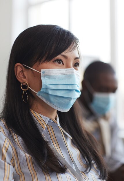 Ritratto verticale di una donna d'affari asiatica che indossa una maschera e guarda lontano mentre è seduta tra il pubblico alla conferenza d'affari