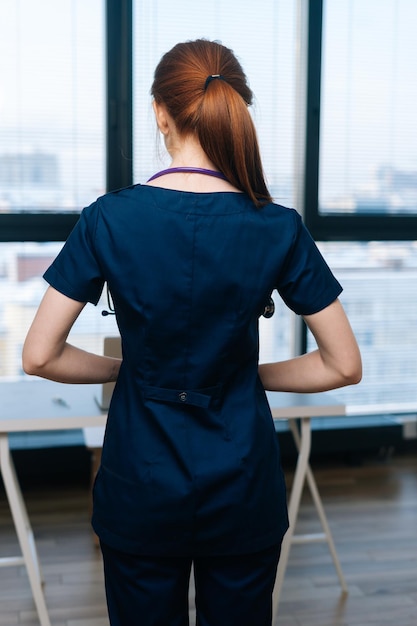 Ritratto verticale di una dottoressa irriconoscibile che indossa l'uniforme medica in piedi sullo sfondo della finestra