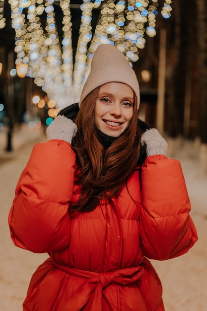 Ritratto verticale di una giovane donna allegra con un cappello e una giacca calda invernale che posa sulla città innevata