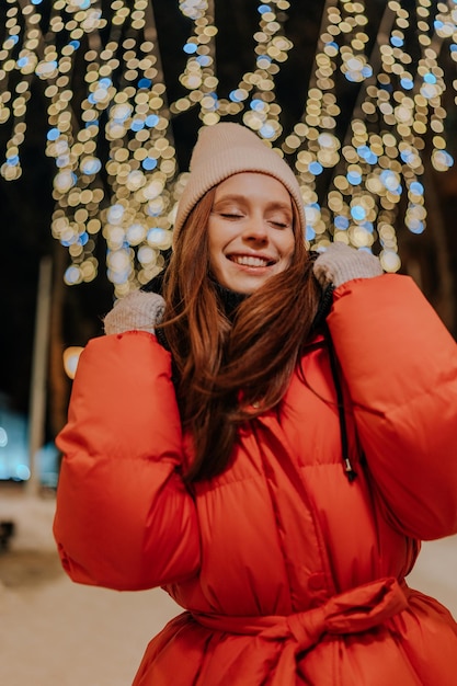 Ritratto verticale di una giovane donna allegra con un cappello e una giacca calda invernale che posa sulla città innevata