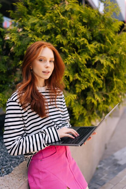 Ritratto verticale di una giovane donna dai capelli rossi in piedi nel parco della città vicino agli alberi con un portatile che scrive usando la connessione WiFi all'aperto guardando la telecamera