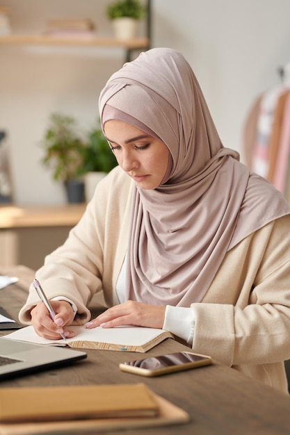 Ritratto verticale di una giovane donna sicura di sé che indossa l'hijab concentrata sul lavoro d'ufficio facendo appunti nel libretto