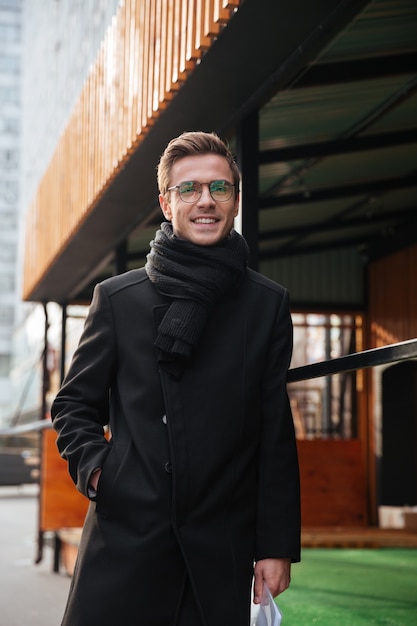 Ritratto verticale di uomo d'affari con gli occhiali e vestiti caldi che cammina per strada con un braccio in tasca