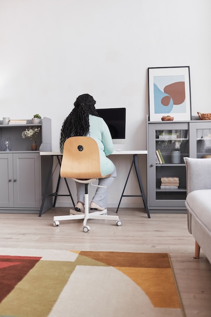 Ritratto verticale di vista posteriore di una donna afroamericana formosa che utilizza il PC sul posto di lavoro di casa in interni minimi, spazio di copia