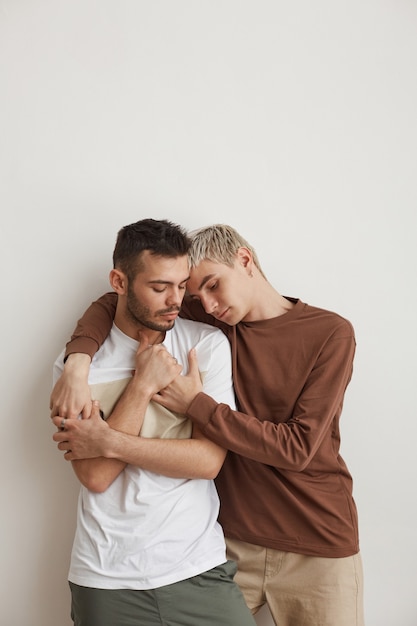 Ritratto verticale in vita di una giovane coppia gay che si abbraccia teneramente mentre si trova contro il muro bianco, toni minimi, spazio copia