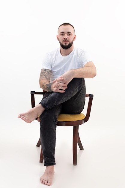 Ritratto verticale tatuato bello brunet fiducioso uomo a piedi nudi barbuto seduto sulla sedia successfu