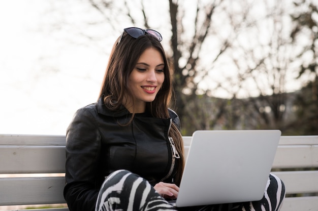 Ritratto vicino della studentessa splendida dei capelli scuri che per mezzo del computer portatile alla città universitaria, adolescente femminile affascinante che studia sul banco di parco con il net-book aperto