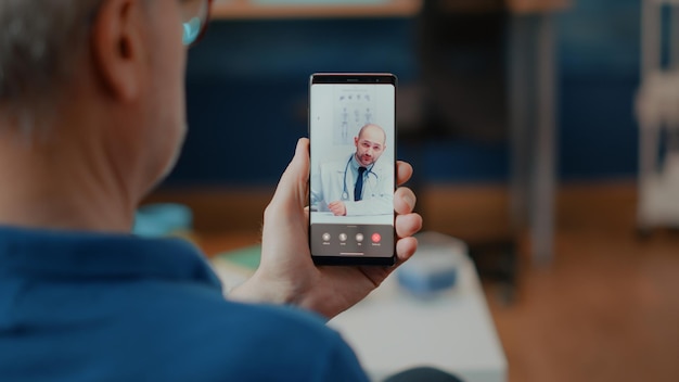 Riunione di una persona anziana in videochiamata online con il medico, che effettua una consultazione medica a distanza. Vecchio che utilizza la teleconferenza sullo smartphone per la telemedicina e la chiamata di telemedicina. Telecomunicazione