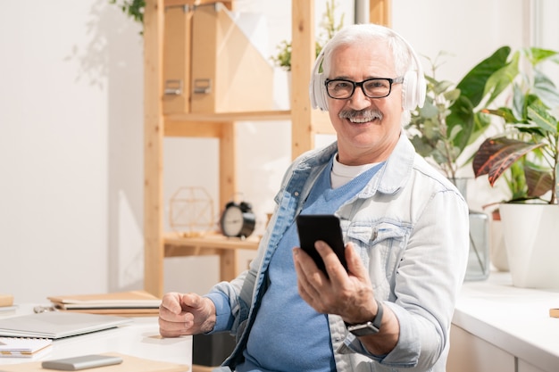 Riuscito uomo invecchiato in cuffie e abbigliamento casual utilizza lo smartphone mentre era seduto alla scrivania