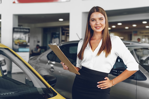 Rivenditore di auto femminile giovane attraente in piedi in showroom vicino a una nuova auto