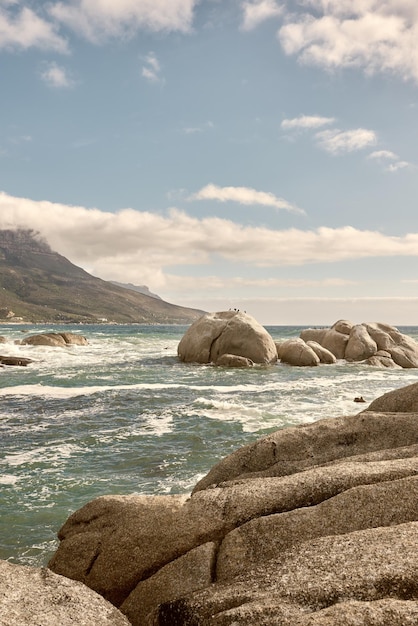 Rocce nell'oceano sotto un cielo nuvoloso blu con spazio per la copia Paesaggio panoramico delle onde della spiaggia che si infrangono contro massi o grandi pietre nel mare in una popolare località estiva di Città del Capo in Sud Africa