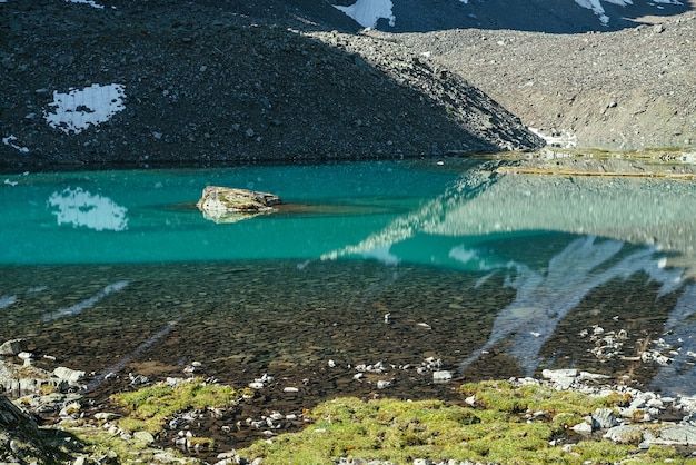 Roccia nel lago di montagna turchese. Montagna innevata riflessa nelle acque azzurre del lago glaciale. Bellissimo sfondo soleggiato con riflesso del ghiacciaio bianco come la neve nella superficie dell'acqua verde del lago di montagna.