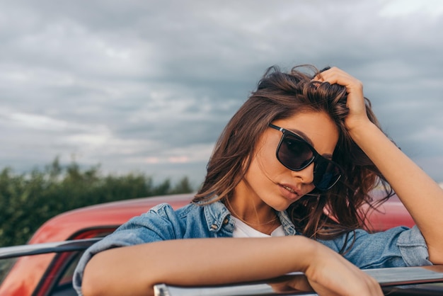 Romantica bella e attraente giovane donna caucasica che indossa occhiali da sole vicino alla sua auto rossa su sfondo nuvoloso Viaggio all'aperto femminile europeo con capelli che soffiano su uno sfondo di campo Concetto di viaggio