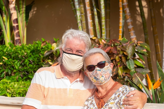 Romantica coppia anziana che si abbraccia in un giardino tropicale indossando una maschera facciale a causa del coronavirus - stile di vita rilassato per due pensionati durante le vacanze estive