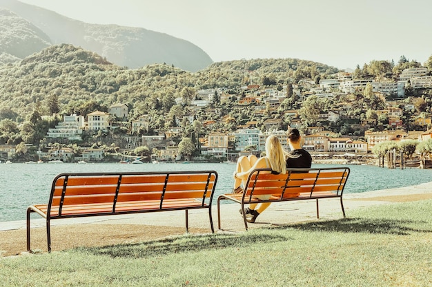 Romantica coppia giovane seduto in panchina ad Ascona località turistica di lusso sul Lago Maggiore nel cantone Ticino in Svizzera in estate. Le persone viaggiano per strada all'aperto. Vacanze costose per famiglie.