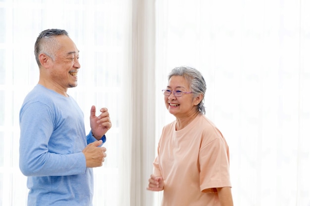 Romantica e allegra coppia senior di benessere asiatico si diverte a ballare e tenersi per mano alla musica insieme a sorrisi e felicità nel soggiorno Attività di pensionamento senior a casa